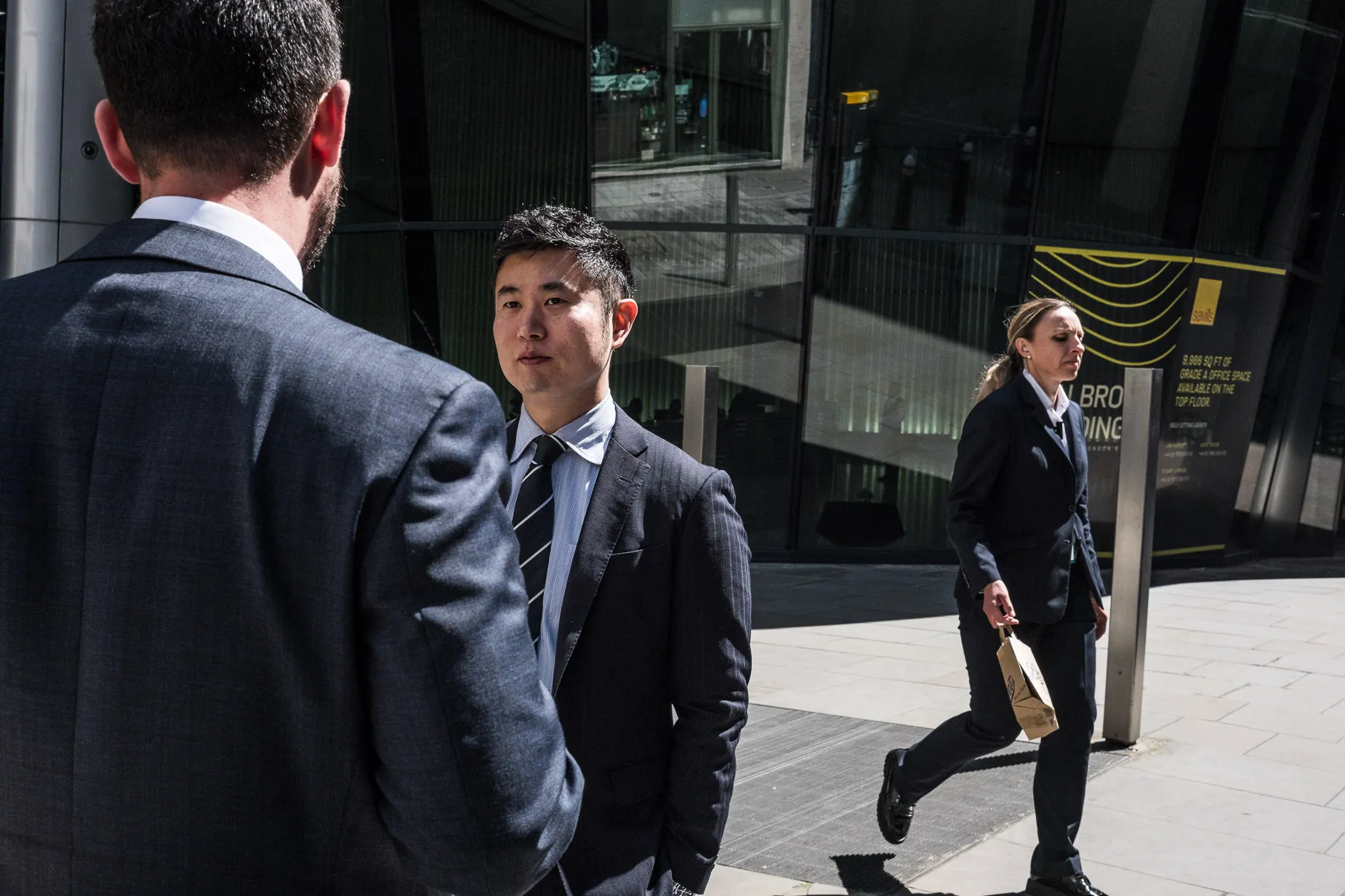 2 suited city men chat, suited city woman walks by in the background