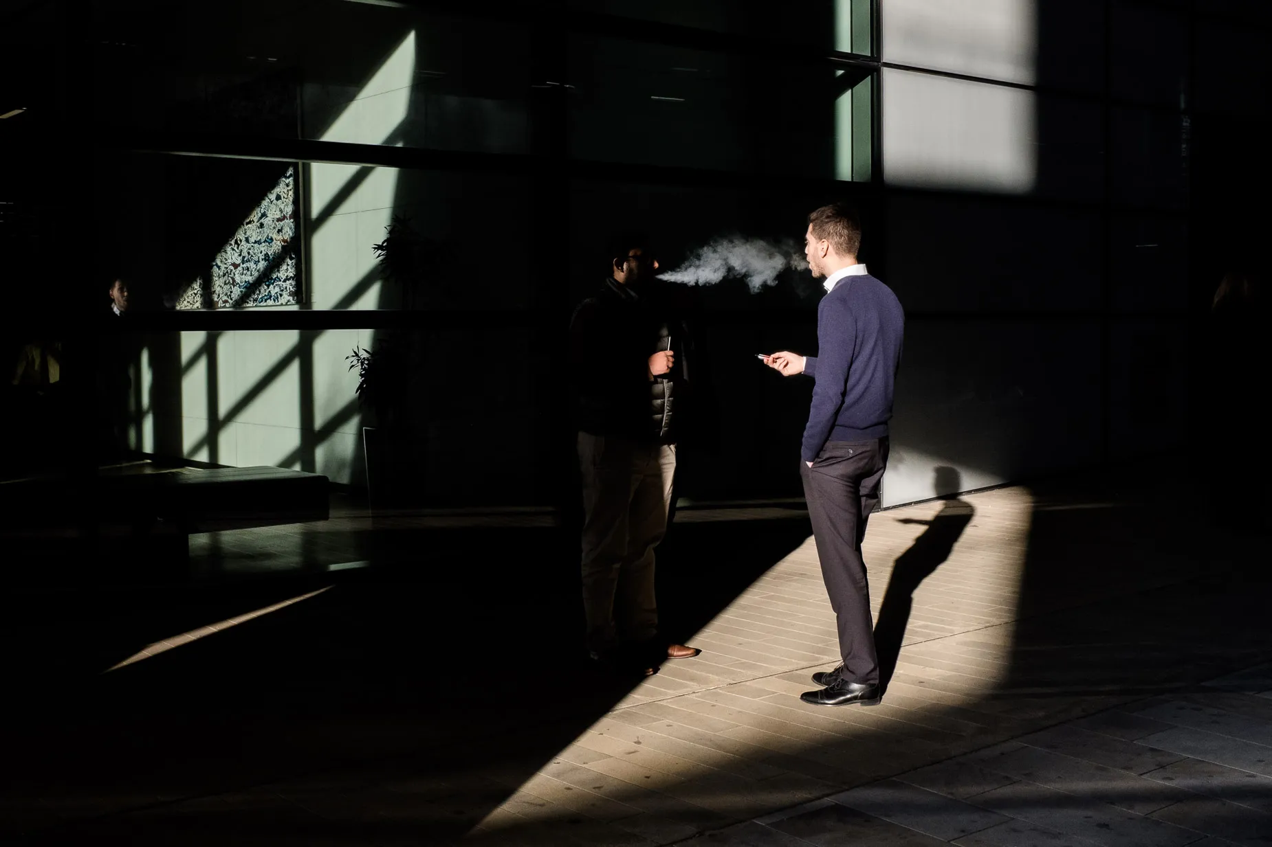 Light and shadow scene of two men smoking, one mans shadow is cast onto the ground